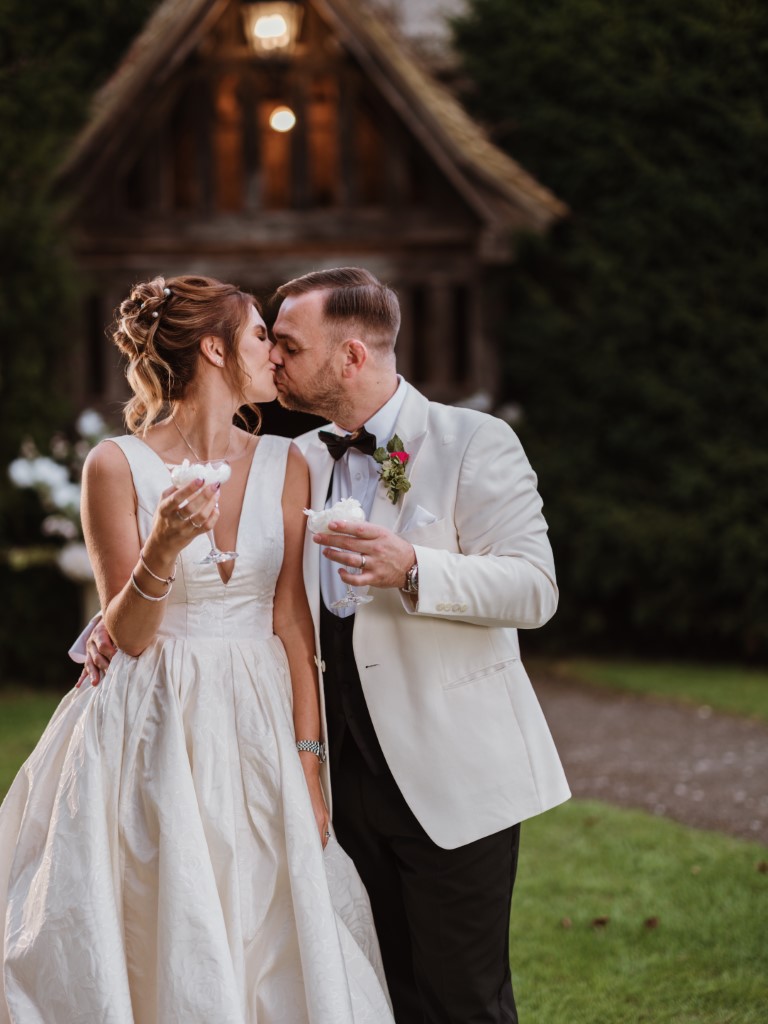 A couple kiss whilst holding confetti served in Champagne Flutes outside the Porch in the evening at Bilsington Priory