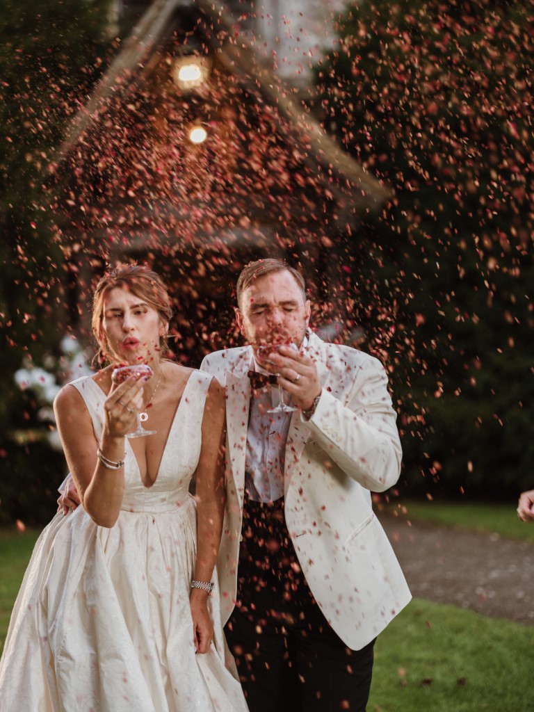 A couple blow confetti towards the photographer outside the porch of Bilsington Priory a wedding venue near ashford